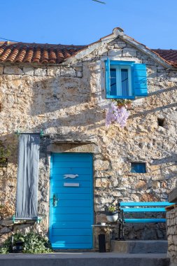Canlı mavi kapıları ve panjurları olan yıpranmış taştan bir ev güneşli bir Akdeniz caddesinde oturuyor. Ivy duvara tırmanıyor, mavi bir bank cazibe katıyor, ve sahne kıyı köyünün sıcaklığını çağrıştırıyor..