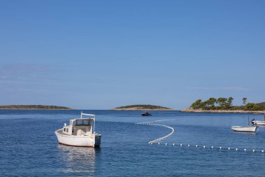 Durgun, berrak bir gökyüzünün altında, şamandıralardan ve uzak adalardan oluşan kıvrımlı bir çizgiyle, sakin mavi sularda süzülen beyaz bir teknenin sakin bir görüntüsü..