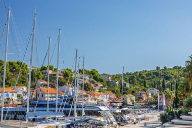 MASLINICA, CROATIA, 08.25.2025: Parlak bir sahil marina sahnesinde açık mavi bir gökyüzü altında demirlemiş sayısız yat ve yelkenli sahnesi yer almaktadır. Sahil boyunca uzanan antika evler, huzurlu, tatil havası yaratıyor..