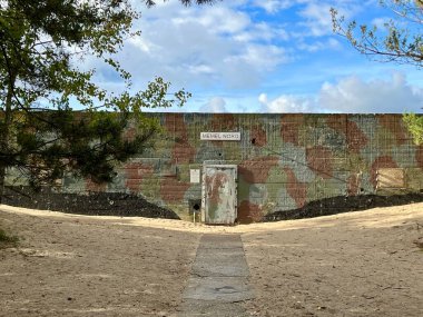Memel-Nord askeri kuvvetleri, Klaipeda, Litvanya. Kumlu arazi ve seyrek bitki örtüsünün ortasında, merkezde yıpranmış, metal bir kapı gösteren kamufle edilmiş bir askeri takviye duvarı bulunuyor.. 
