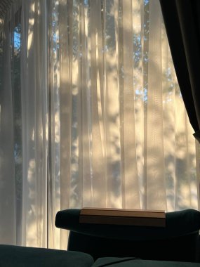 Yaprakların yumuşak gölgeleri pencereden sarkan beyaz perdelere dökülür. Gölgeler doğal bir desen oluşturuyor. Bu da güneş ışığının yakınlardaki yaprakların arasından süzüldüğünü gösteriyor.. 