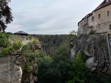 Almanya, Saksonya 'daki Ortaçağ Hohnstein Kalesi. Taş kale, aşağıdaki ağaçlık vadiyle kayalık bir uçurumun tepesine tünemişti. Yıpranmış duvarları ve geleneksel kırmızı kiremitli çatısı olan tarihi yapı..
