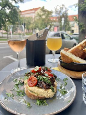 İki kişilik romantik bir yemek. Küçük bir açık hava masasında, içinde humus, domates ve otlar bulunan gurme açık bir sandviç gri bir tabakta sunulur, maydanozla süslenir. Bir bardak şarap ve hafif bir bira. 