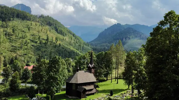 Slovakya 'nın Tatranska Javorina kentindeki kilisenin ve Tatra dağlarının çarpıcı panoramik manzarası. Yoğun orman ve yuvarlanan tepelerle çevrili yemyeşil vadide yuva yapmış ahşap kilise.