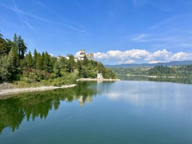 Polonya 'daki Niedzica Kalesi' nin panoramik manzarası. Kale yemyeşil bir yer ve sakin bir göl ile çevrili bir tepeye tünemişti. Ortaçağ yapısında taş duvarlar ve kuleler yer alıyor.