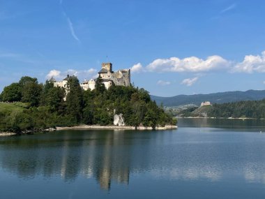 Polonya 'daki Niedzica Kalesi' nin panoramik manzarası. Kale yemyeşil bir yer ve sakin bir göl ile çevrili bir tepeye tünemişti. Ortaçağ yapısında taş duvarlar ve kuleler yer alıyor.