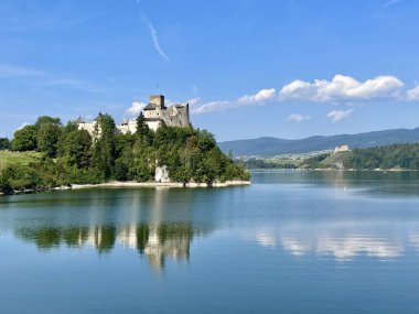 Polonya 'daki Niedzica Kalesi' nin panoramik manzarası. Kale yemyeşil bir yer ve sakin bir göl ile çevrili bir tepeye tünemişti. Ortaçağ yapısında taş duvarlar ve kuleler yer alıyor.