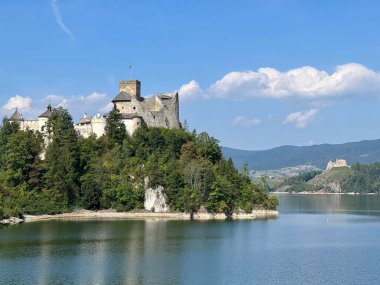 Polonya 'daki Niedzica Kalesi' nin panoramik manzarası. Kale yemyeşil bir yer ve sakin bir göl ile çevrili bir tepeye tünemişti. Ortaçağ yapısında taş duvarlar ve kuleler yer alıyor.