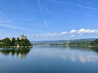 Polonya 'daki Niedzica Kalesi' nin panoramik manzarası. Kale yemyeşil bir yer ve sakin bir göl ile çevrili bir tepeye tünemişti. Ortaçağ yapısında taş duvarlar ve kuleler yer alıyor.