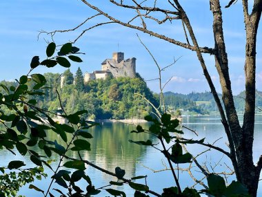 Polonya 'daki Niedzica Kalesi' nin panoramik manzarası. Kale yemyeşil bir yer ve sakin bir göl ile çevrili bir tepeye tünemişti. Ortaçağ yapısında taş duvarlar ve kuleler yer alıyor.
