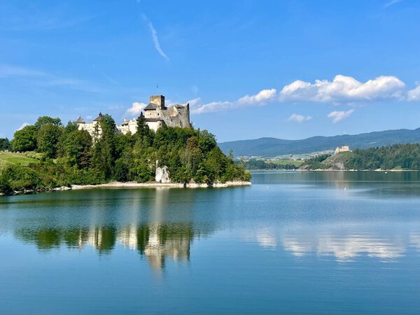 Panoramic view of Niedzica Castle in Poland. The castle perched on a hill surrounded by lush greenery and a serene lake. The medieval structure features stone walls and towers