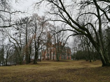 Lentvaris Malikânesi Parkı, Litvanya. Kırmızı tuğlalı ve sivri çatılı tarihi bir bina yapraksız ağaçlarla kısmen gizleniyor..