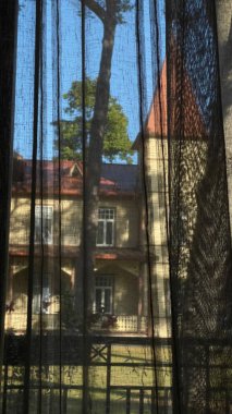 Kapkara perdeleri kısmen açık olan uzun pencere tarihi bir binayı gösteriyor. Binanın sivri kırmızı çatısı, bej duvarları ve simetrik beyaz çerçeveli pencereleri vardır.