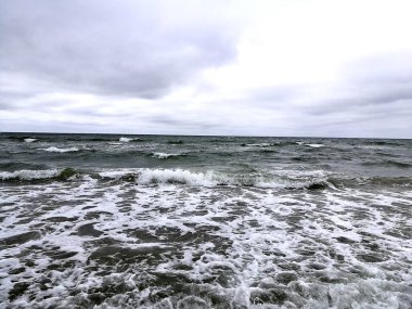 Baltık Denizi 'nin dalgaları bulutlu ve bulutlu bir gökyüzünün altında kıyıya doğru yuvarlanıyor. Dalgalı su, kumlu sığ sulara çarptığında beyaz köpük oluşturur.. 