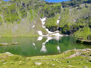 Mountain Lake Balea, Romanya. Dik, kayalık yamaçlara kurulmuş berrak yeşil sular kısmen yemyeşil çimenler ve kar parçalarıyla kaplı. Göl çevresindeki manzarayı yansıtır.. 