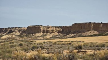 Navarre, İspanya 'daki yarı çöl Bardenas Reales' in panoramik manzarası. Engebeli, aşınmış kayalıkların berrak bir gökyüzüne karşı kurulduğu çorak bir manzara. Nadir bitki örtüsü toprağı kaplar