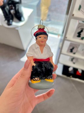 Geleneksel bir Katalan Caganer heykelciğine benzeyen küçük bir heykelcikle el ele tutuşmak. Karakter tipik bir şekilde giyinmiştir: beyaz gömlek, siyah pantolon ve kırmızı şapka.. 