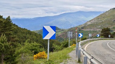 Dağlardaki yolun panoramik manzarası. Arabayla yolculuk. Metal korkulukları ve mavi ok trafik işaretleriyle dolambaçlı dağ yolu. Yol kenarındaki yemyeşil bitki örtüsü sarı çiçekli.