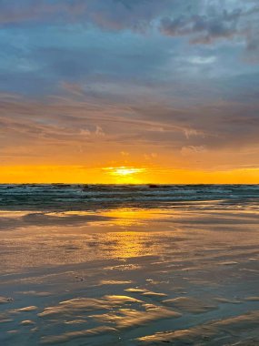 Letonya, Liepaja 'da Baltık Denizi üzerinde gün batımı manzarası. Turuncu güneş kısmen ufkun üzerindedir, gökyüzünde sıcak bir parıltı yayar ve nazik dalgaları yansıtır. Huzur ve doğal güzellik