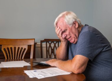 Gri tişört giyen beyaz saçlı yaşlı adam faturaları öderken stresli görünüyor.