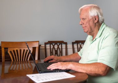 Beyaz saçlı ve yeşil tişörtlü yaşlı bir adam yemek odasındaki derme çatma bir ofiste dizüstü bilgisayarında çalışıyor..