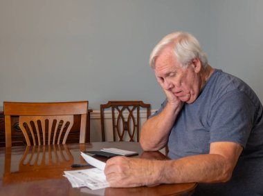 Gri tişört giyen beyaz saçlı yaşlı adam faturaları öderken stresli görünüyor.