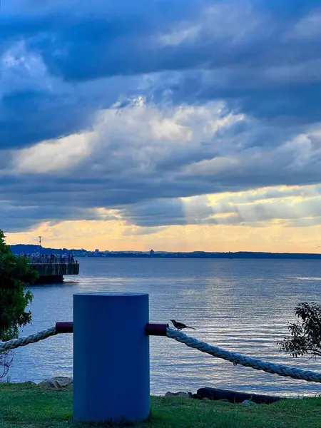 Alacakaranlıkta çekilen sakin bir göl manzarası dramatik bulutlar ve yumuşak güneş ışığıyla dolu kasvetli bir gökyüzünü gösteriyor. Ön planda, kalın ipli sağlam bir boğa gözü rengini yansıtan sakin suya götürür.