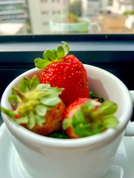 Yeşil yapraklı taze çilekler içeren beyaz bir kupa, bitkilerle süslenmiş, bulanık şehir manzarası sunan bir pencerenin yanına yerleştirilmiş, tazelik ve hafiflik hissi iletiyor.