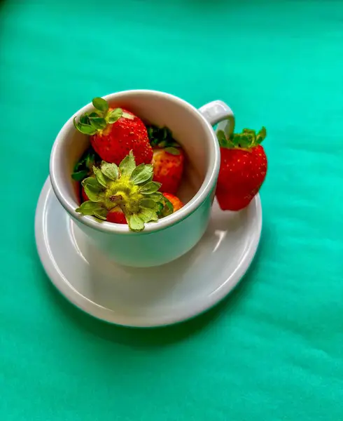 Taze, sulu çileklerle dolu beyaz bir kupa ve yanında da bir dilim çilek. Canlı yeşil arka plan çileklerin kırmızı rengini vurguluyor, neşeli ve iştah açıcı bir sahne yaratıyor..