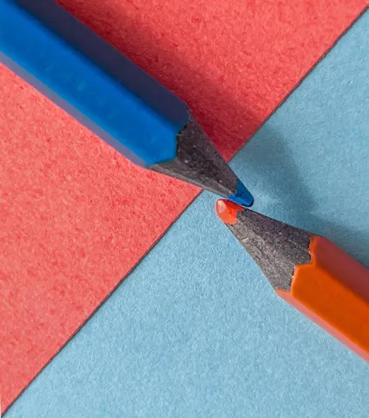 İki renkli kalem, biri mavi ve biri turuncu, kırmızı ve mavi kağıttan oluşan canlı bir arka planın ortasında buluşuyorlar. Görüntü yaratıcılığı ve işçiliği çağrıştırıyor, minimalist ve neşeli bir ortamda keskin renkli uçları vurguluyor..