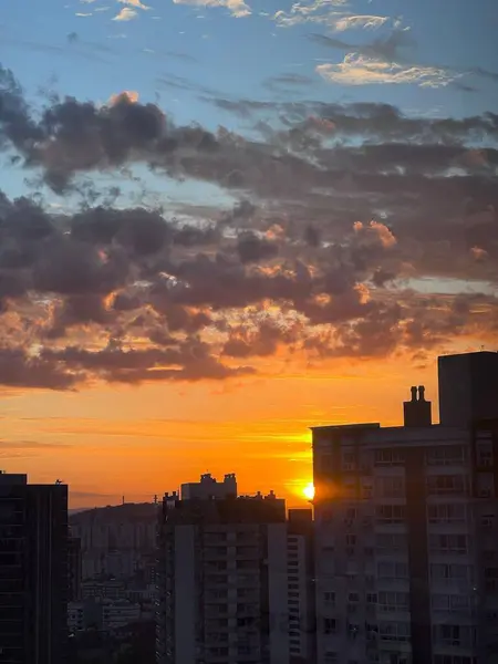 Renkli bulutların sıcak turuncu ve sarı renkleri yansıttığı modern bir şehirde güzel bir gün batımı. Ön plandaki yüksek binalar dramatik gökyüzü ile tezat oluşturarak sakin ve davetkar bir atmosfer yaratıyor..