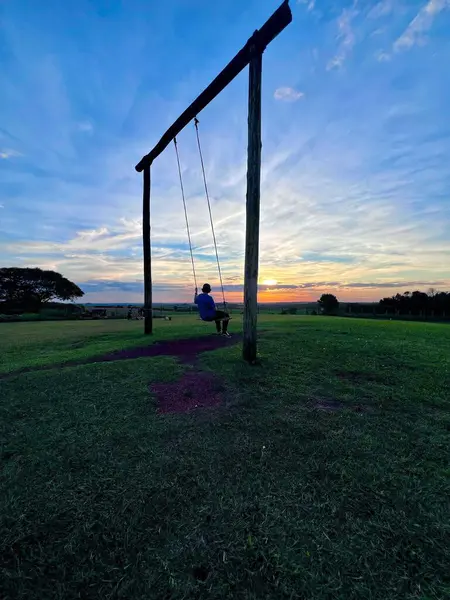 Bir çocuk ahşap bir salıncakta oturur ve kırsalda güzel bir günbatımının tadını çıkarır. Gökyüzü mavi ve turuncu tonlarıyla patlar, sakin ve yansıtıcı bir sahne yaratır. Salıncak, yemyeşil yemyeşil, güçlü bir ahşap çerçeveye sarkıyor.