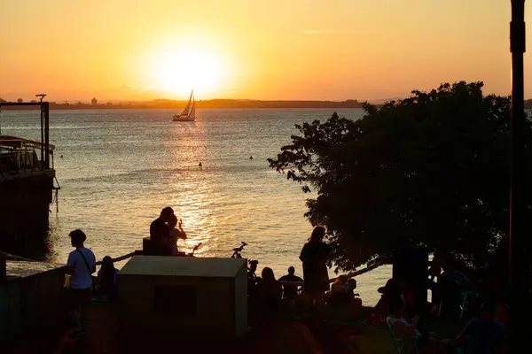 Deniz kenarında sakin bir günbatımı sahnesi, açık havada eğlenen insanların siluetleri. Ufukta süzülen bir yelkenli batan güneşin altın ışığını yansıtıyor. Atmosfer rahatlamayı ve doğayla bağlantıyı iletir..