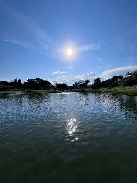 Açık bir günde güneşi yansıtan sakin bir göl manzarası, arka planda bereketli bir manzara. Sakin su, mavi gökyüzü ve beyaz bulutlarla çelişiyor. Huzurlu ve rahatlatıcı bir atmosfer yaratıyor. İnsanlar güneşli havalarda parktan hoşlanırlar.