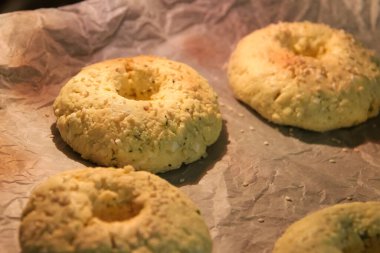 Ev yapımı peynirli simitlerin fırının içinde pişirilme sürecine yakın çekim görüntüsü. Hamur halka şeklinde şekillendiriliyor ve üzerine susam tohumları serpiştiriliyor, parşömen kağıdının üzerine konuyor. Şey...