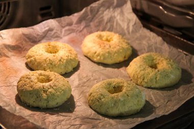 Ev yapımı peynirli simitlerin fırının içinde pişirilme sürecine yakın çekim görüntüsü. Hamur halka şeklinde şekillendiriliyor ve üzerine susam tohumları serpiştiriliyor, parşömen kağıdının üzerine konuyor. Şey...