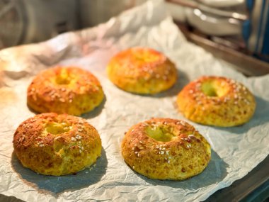 Ev yapımı peynirli simitlerin fırının içinde pişirilme sürecine yakın çekim görüntüsü. Hamur halka şeklinde şekillendiriliyor ve üzerine susam tohumları serpiştiriliyor, parşömen kağıdının üzerine konuyor. Şey...