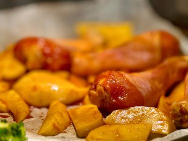 İçinde fırın tepsisi olan açık bir fırının yakın plan fotoğrafı, altın patates dilimlerinden yapılmış bir yatakta salamura edilmiş tavuk budu ve yanında da bir sıra canlı yeşil brokoli çiçekleri. Şey...