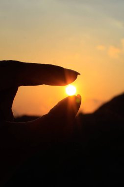 Gün batımında güneşi tutan bir el. Görüntüde, bir elin silueti canlı bir günbatımı gökyüzüne karşı siluetlenir, güneş başparmak ve işaret parmağı arasına yerleştirilir, elin onu tuttuğu izlenimini verir.. 