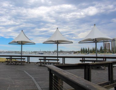 Üç şemsiye şeklindeki tente hattı ahşap bir rıhtım geçidi, ziyaretçilere tekne dolu marinaya ve bulutlu bir gökyüzünün altındaki ufuk çizgisine hayran oldukları için gölge sunuyor..