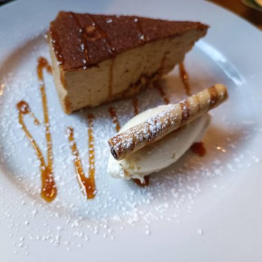 Lezzetli bir kremalı karamel peynirli kek, bir tutam krem şanti ve rulo gofretle süslenmiş, beyaz bir tabakta zarif bir şekilde karamel sosuyla sunuluyor..