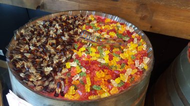 Kola ve meyve aromalı renkli jelibon çeşitleri, ahşap bir fıçıda sergileniyor, şeffaf plastikle kaplı, self servis için metal bir sapı var..