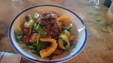 Canlı ve taze bir gurme domuz salatası mavi kenarlı beyaz bir kasede servis edilir, kiraz domatesleri, roka, ızgara kabak ve ananas parçaları rustik ahşap bir masa üzerinde..