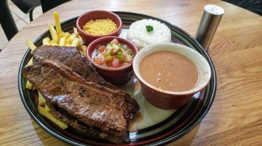 Yanında taze domates sosu, kremalı pilav, geleneksel Brezilya farofası ve ahşap bir masada servis edilen bir kase kahverengi fasulye ile lezzetli ızgara biftek ve kızarmış patates..