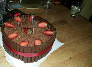 Çikolatalı pasta üzerine taze çilek, çikolata çubukları ve kırmızı kurdeleyle çevrili, ahşap bir masaya yerleştirilmiş, neşeli ve hoşgörülü bir kutlama atmosferi yakalıyor..