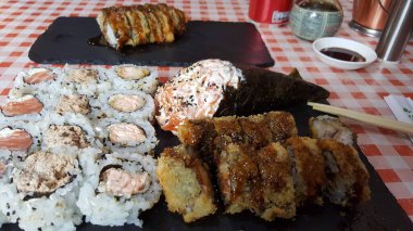 Somon ruloları, temaki ruloları ve kareli masa örtüsünde servis edilen siyah bir tabakta servis edilen kızarmış suşi de dahil olmak üzere çeşitli suşiler..