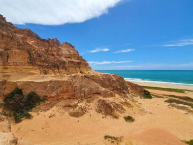 Engebeli uçurumları olan kayalık bir kıyı manzarası, berrak mavi gökyüzü ve arka planda sakin bir turkuaz okyanus, doğanın sakin güzelliğini resmediyor..
