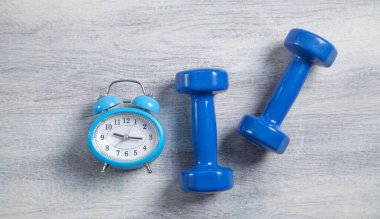 Beyaz ahşap arka planda mavi halterler ve çalar saat.