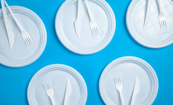 White plastic plates, forks and knifes on blue background.