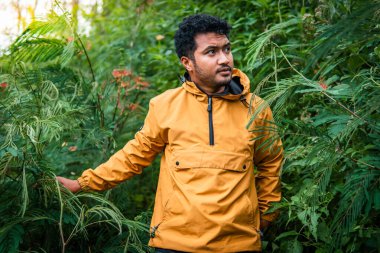 Sarı ceket giyen bir adam yoğun bitki örtüsü ve tropikal bitkilerle çevrili yemyeşil bir ormanı keşfeder. Güneş ışığı ağaçların arasından süzülüp sakin ve maceracı bir atmosfer yaratır..